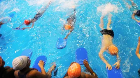 PA Media A group of children use buoyancy aids to paddle through a swimming pool. They have red, white or orange swimming caps. There is also a line of children at the edge of the pool after they finished their paddle.