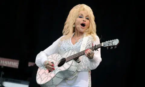 Dolly Parton wearing a white and glittering costume plays a guitar and sings on stage while smiling