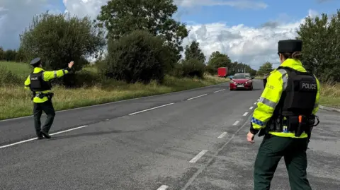 Two police officers are standing at opposite sides of a main road. They are wearing full uniform including hats, high visibility jackets and padded vests. 