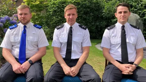 BBC PCSO TIM Parry, Sgt Greg Gillespie and PC Luke Holden sit on chairs in a garden. They all wear a police uniform. PCSO Parry wears a blue tie and libraries on his shoulders. Sgt Gillespie and PC Holden wear black relationships and book on their shoulders.