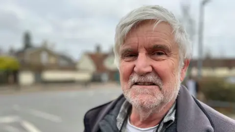 JO THEWLIS/BBC Brian Ross has short white hair and a short white beard.
He is wearing a grey fleeced jacket with a grey jumper underneath. He is smiling at the camera with buildings and a road behind him.