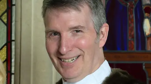 A man with short greying hair wearing a white shirt and a red ceremonial coat. 