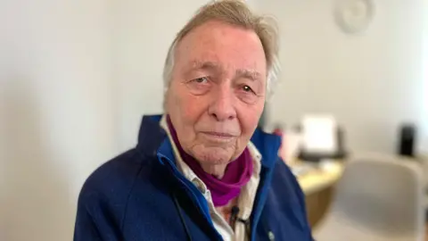 BBC/Martin Giles Ian Miller looks directly at the camera. He is wearing a navy fleece and has grey hair with has some light brown to it at the front. The light has lit his pupils. He has a cerise pink scarf around his neck and a shirt underneath his fleece. The surgery room is visible but blurred behind him which is modern and white. He looks serious.