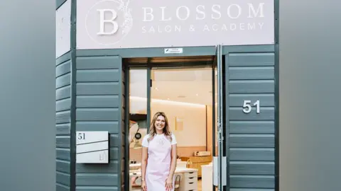 Lucy Rebecca Photography A woman with blonde and brown hair wearing a white t-shirt and a pink apron is standing outside a building with dark grey wood panels. The number 51 is on the outside, and the sign above the door is pink with white writing that reads "Blossom Salon and Academy"
