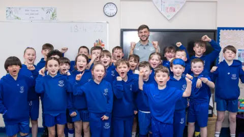 Everton Group of children in blue kits stand in a class room with Everton football player Kiernan Dewsbury-Hall 