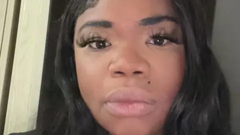 Stacey Llewellyn poses for the camera. She is a young black woman with long hair and false eyelashes.