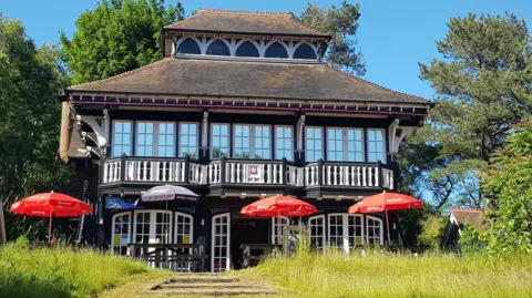 A Victorian sports pavilion with large windows and balconies is in view with trees and blue skies around it and parasols in the foreground covering picnic tables