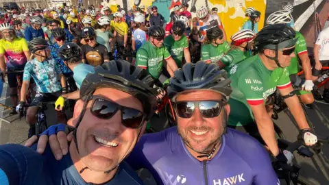 Graham Bell Graham smiles as he takes a selfie with a friend and they're at the start of a bike race with numerous other participants in the background, they're wearing helmets and sunglasses