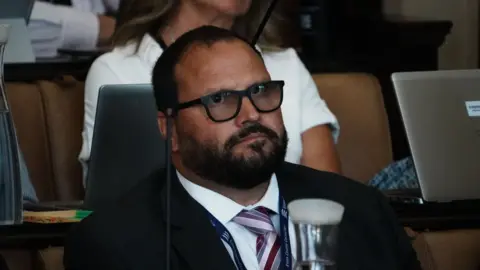 Chris Hossack is wearing glasses and has short black hair and a beard. He is wearing a suit while sitting in the council chamber.