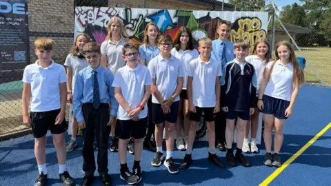 The photo shows thirteen students all looking at the camera. Some are wearing PE kits, others are wearing uniform. They're stood on a bright blue tennis court. 