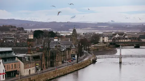 Getty Images A general view of Inverness with lots of birds in the sky. The river is on the right, with a bridge crossing it. There are buildings and a road on the left. Hills are visible in the distance. 