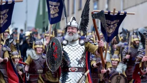 PA Media The guizer jarl in full Viking dress. He is holding a shield with a bird insignia and a helmet with metallic wings on the side. He holds aloft an axe. He has a very full grey beard. Behind are more men dressed in Viking costume.  