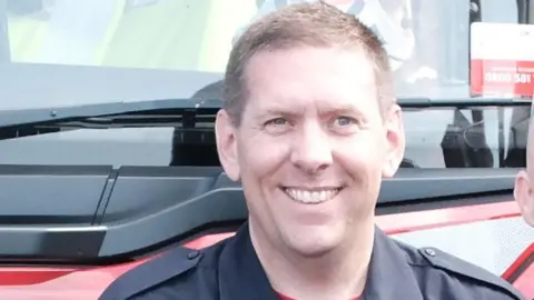 A smiling man with short brown hair and a dark blue shirt stands in front of a red vehicle.