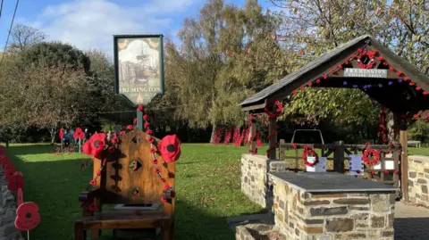 Fremington Village Green decorated for Armistice Day. A central area features a memorial setup with red poppies. There is a large wooden chair adorned with poppies; as is a shelter, which also has garlands.