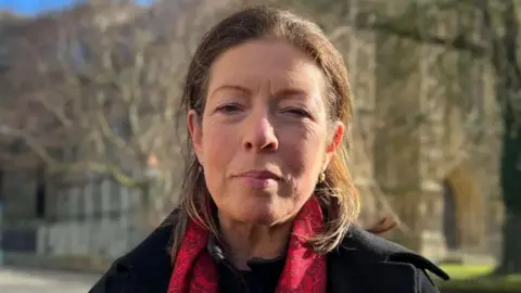 BBC/David Reeves A head and shoulder shot of a woman with dark eyes and shoulder-length brown hair. She is wearing a black coat and a red and black scarf. She is stood in front of Beverley Minster with the background out of focus.