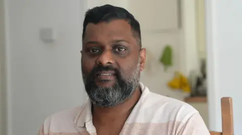 Sumesh has a dark beard and is wearing a beige polo shirt. He is sitting in a wooden chair and smiling at the camera