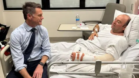 Chris Minns/Facebook A picture inside a hospital room. On a grey hospital bed with white sheets lies Mr Ahmed, a balding man wearing a white t-shirt with his left arm and wrist in a cast. Chris Minns sits at the end of the bed smiling at him, wearing a light blue shirt, dark blue tie and black trousers.
