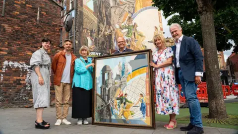 Simon Dewhurst Six people stand around a framed picture which is replicated as a mural on the wall behind them. The picture is called Cornucopia and is of a man with some corn against the backdrop of some iconic Leeds buildings. The goddess Cornucopia is also on there.