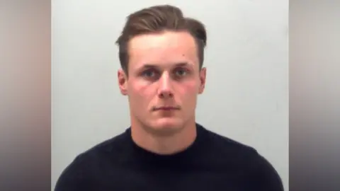 The head and shoulders of a man with brown hair slicked back, stands against a white background, wearing a black T-shirt. He is looking at the camera.