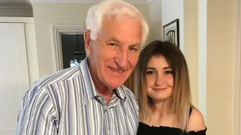 Charlotte Andrews Richard, an older man with white hair wearing a striped shirt stands beside his daughter Charlotte, a younger woman with long hair in a black off-shoulder top. The background includes a kitchen and dining area.