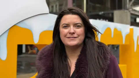 Martina Connolly stands in front of a large, yellow sign with 'Belfast'. She is a young woman with large, dark hair and she is wearing a purple coat with a fur trim collar. She is smiling at the camera.