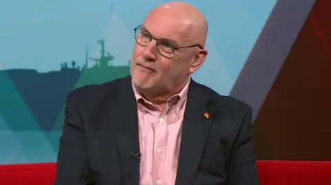 Nick Adams-King is bald and wearing thin circular glasses. He has a dark blazer on over a pink shirt. He is sitting on a red sofa with a red and blue wall of graphics behind him.