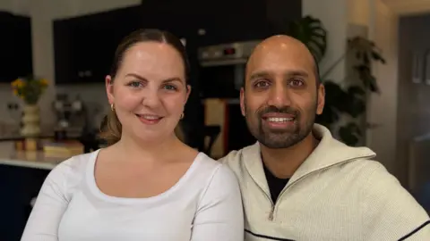 Ben Schofield/BBC Hannah and Asantha sitting next to each other and looking directly down the camera. Both are smiling and showing their teeth. They are sitting inside, with a kitchen area behind them. Hannah is wearing a white, round-necked top, and has two earrings hanging from her ears. Her hair is tied back in a ponytail, which we can only see a hint of behind her. Asantha appears to be bald on the top of his head and has dark stubble across the lower half of his face. He is wearing a cream half-zip top with a collar that is open and laying over his shoulders. 