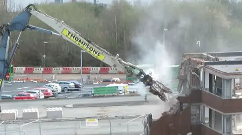 Gateshead Council The arm of a crane with pincer-like claw is biting at a building to bring it down. There are cars parked beyond and fencing round the area being demolished. The crane says Thompsons. The building is half destroyed.