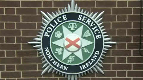 A crest from the Police Service of Northern Ireland. It is round with a green centre with a number of symbols on it and the words Police Service of Northern Ireland in white writing on a black border.