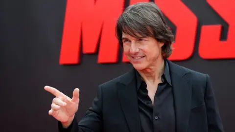 Reuters Tom Cruise pointing off camera with a large red MissionL Impossible sign behind him at the film's premiere