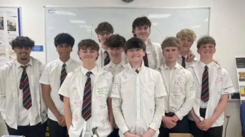 Ten schoolboys are standing in a classroom wearing white shirts that have been signed with colourful pens. Seven of them are wearing maroon and navy striped ties. There is a wall-mounted whiteboard in the background.