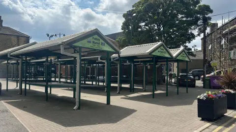 Empty market stalls painted green in Pudsey Market Square on a sunny day.