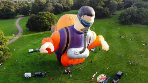 A large orange and purple Action Man hot air balloon is inflated in a grassy area surrounded by spectators and trees.