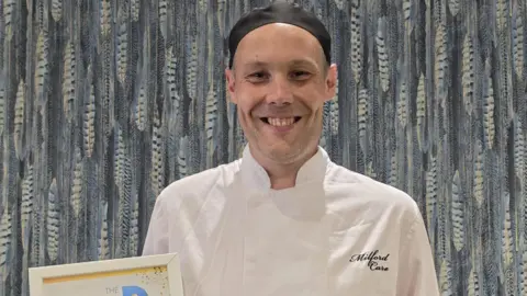 Supplied Glover holding a plaque while in his chef uniform. 