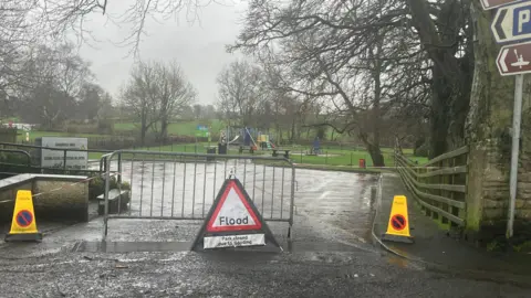 A flood sign in front of an entrance to a park. It reads "Park closed due to flooding". Beside it are two traffic cones.