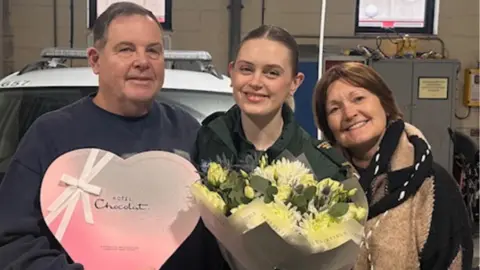 Simon Thompson is on the left and has short brown hair. He is wearing a navy jumper and is holding a heart shaped box of chocolates. Chloe Spencer is in the middle and has her hair in a ponytail. She is holding a bunch of flowers. Tina Thompson is on the right and has short brown hair and is wearing a black and beige coat.