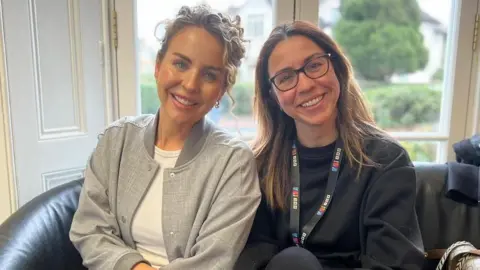 BBC Essex Lydia Bright sits on the right-hand side of a black sofa next to Sonia Watson. Lydia wears a light grey jacket with white top underneath. She has curly blonde hair which is tied back. Sonia is wearing a black jumper with a black BBC lanyard around her neck. She has brown hair below her shoulders and she has dark-rimmed glasses on. Both are smiling at the camera. They sit on a sofa in front of a window which has white wood panelling surrounding it, and blurred trees can be seen out of the window.