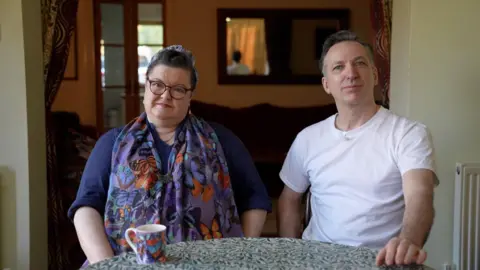 Maggie and Jarvis sit at a kitchen table, she wearing a purple dress and matching scarf, him a white t-shirt. A coffee cup is on the table in front of them.