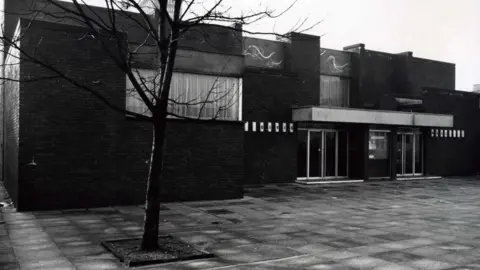 Rochdale Council A black and white photograph of Heywood Civic before the building was refurbished. The image shows the 1960s building, a public square and a tree.