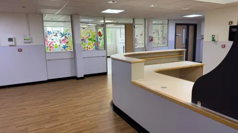 Queen Elizabeth Hospital The inside of a hospital ward. The lower half of the walls and desk where staff would sit are painted lilac. There are internal windows with painted flowers on them. It is empty and the floors are laminated wood.