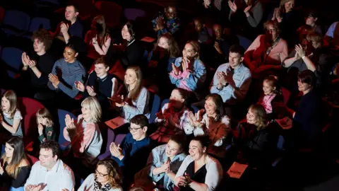 Frasershot Studios Audiences at Royal & Derngate, most of them are clapping their hands and smiling. A mixture of ages are in the seats, they are all looking towards a stage, that cannot be seen. 