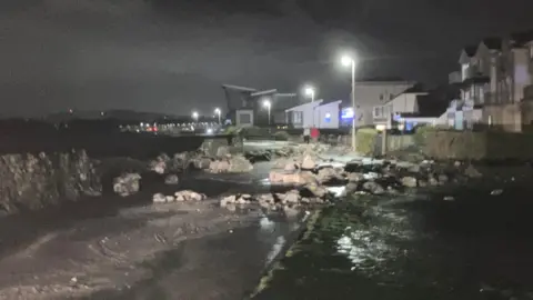 A number of large rocks near the seawall are visible scattered along a footpath. A number of street lights and buildings can be seen in the background