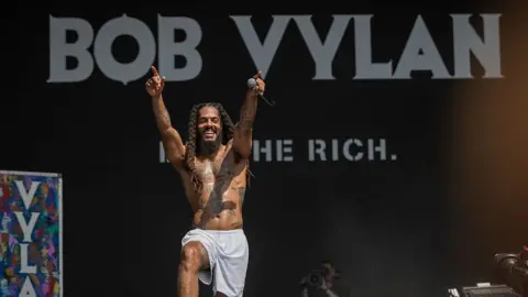 Getty Images  Bob Vylan performs on the West Holts stage during day four of Glastonbury Festival 2025. He is shirtless, wearing white shorts, and holding a mic in his left hand. The stage backdrop is black with white lettering.