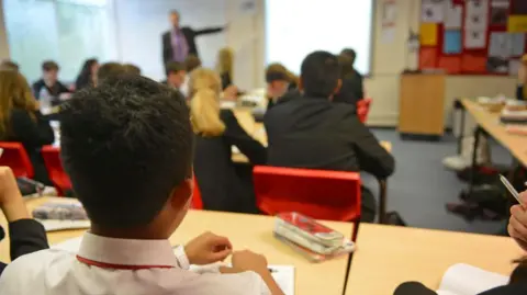 A classroom with students dressed in school uniforms, seated at desks. The students appear engaged in a lesson, facing a teacher at the front of the room who is gesturing towards a presentation screen. The desks have stationery, including pencil cases, notebooks and other study materials.