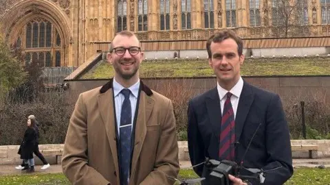 Local Democracy Reporting Service (LDRS) Image of two men standing outside the Houses of Parliament in London. One man is wearing a camel overcoat, a tie and round glasses, The other man is dressed in a black suit and holding a drone.
