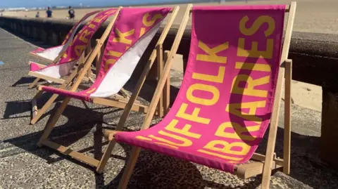 A pink deck chair which says Suffolk Libraries on it in yellow is on the promenade in Lowestoft. You can see blue skies and the beach in the background. 