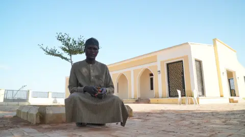 Gift Ufuoma / BBC Masud Abdulrasheed, sitting outside the new mosque in the village