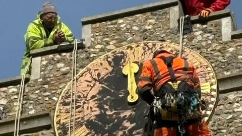 Reverend Daniel Whiffin A worker wearing an orange hi-vis jacket and a hard hat dangling next to the top of St Mary's Church in front of a large clock. Another worker, wearing a yellow hi-vis hacket is watching on from the top of the tower. 