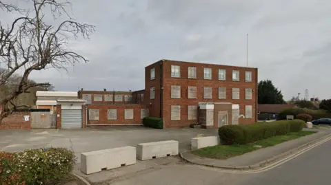 A two-storey flat-roofed brown brick building. In front of the building is an empty grey car park and hedgerow. The sky is grey