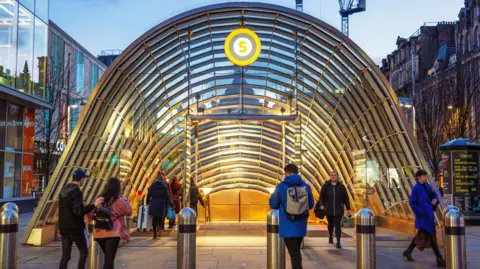 Getty Images The entrance to a subway station. There is a large glass archway with people entering and leaving the station and the Glasgow subway logo at the top of the arch.
